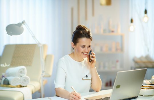 Happy,Woman,Worker,With,Laptop,Speaking,On,A,Smartphone,In